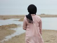Kvinde i lyserød jakke med sort tørklæde om hovedet med ryggen til på stranden